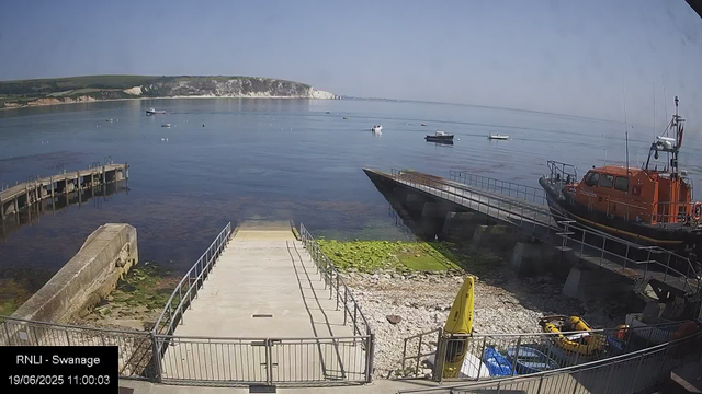 A view of a small harbor with a concrete pier leading out into calm water. To the right, a bright orange rescue boat is stationed on a dry dock with a docked smaller boat nearby. Several boats are floating on the water in the distance, with a large chalk cliff formation and greenery on the shoreline beyond. The sky is clear with no clouds. In the bottom left corner, there is a timestamp and location label indicating "RNLI - Swannage" with the date and time "19/06/2025 11:00:03".