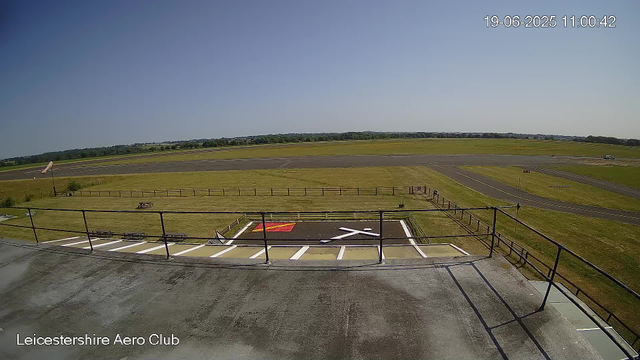A view from a webcam at Leicesterhire Aero Club showing an empty observation deck with a parking lot below. In the background, there is a grassy airfield with a few taxiways and a parked vehicle. The sky is clear with no clouds, and the scene is brightly lit, indicating a sunny day. The timestamp on the image reads 19-06-2025 11:00:42.