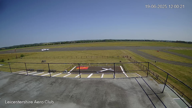 A view from a webcam at Leicesterhire Aero Club showing an empty tarmac area with a small aircraft parked on the runway. There are grassy fields surrounding the runway, with a clear blue sky overhead. The date and time displayed in the top right corner are 19-06-2025 at 12:00:21. The photo includes a railing in the foreground and a parking space area labeled "X" with yellow and red markings.