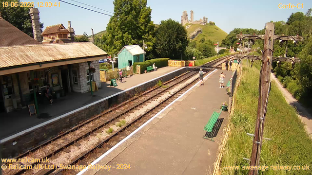 A small train station platform with a few people waiting. There are green benches, a green shed, and some potted plants. A railway line runs through the middle of the scene, with overhead power lines supported by wooden poles on the right side. In the background, a grassy hill with a castle ruin on top is visible, along with trees and other buildings. The sky is clear and sunny.