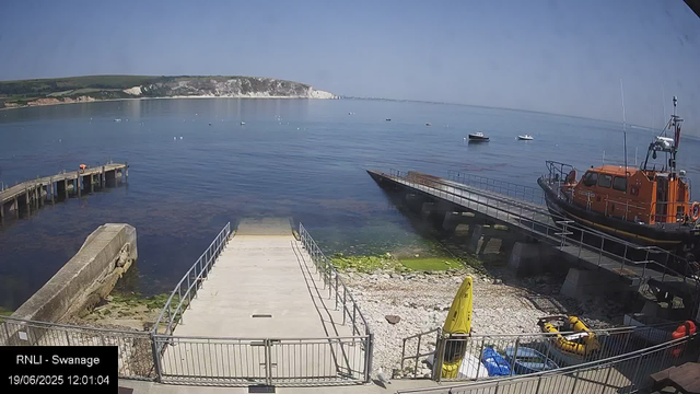 A view of a harbor with a small boat ramp leading into the water. To the right, there is an orange rescue or patrol boat on a slipway. Several small boats are anchored in the calm water, with a distant shoreline featuring white cliffs and green hills under a clear sky. A yellow life raft and some equipment are visible on the rocky area near the ramp. The scene is captured during the day, as indicated by the timestamp.
