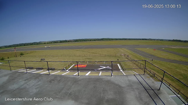 A view of an airport runway and taxiway from a webcam at Leicestershire Aero Club. The image shows a small airplane on the runway, a clear blue sky, and a grassy area surrounding the tarmac. The timestamp in the upper right corner indicates it was taken on June 19, 2025, at 13:00:13.