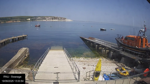 A calm harbor scene with a concrete pier extending into the water, flanked by metal railings. To the right, there is a bright orange rescue boat on a ramp, with yellow and black inflatable rescue boats nearby. Several small boats are anchored in the water, and in the background, the coast with cliffs and greenery stretches along the horizon under a clear blue sky.