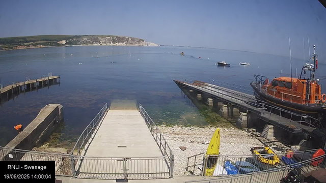 A view of a harbor with calm water, several small boats floating on the surface, and a large orange rescue boat docked on the right. There are piers extending into the water, and people are visible near the lower left corner. In the background, cliffs and greenery line the distant coastline under a clear blue sky. The timestamp in the lower left indicates the date and time as 19/06/2025 at 14:00:04.