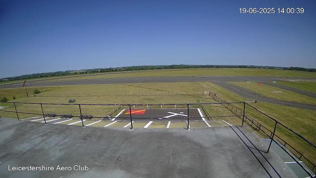 A view from a webcam at Leicesterhire Aero Club showing an empty tarmac with a runway in the distance under a bright blue, clear sky. The foreground features a parking area with white painted lines and a disabled parking symbol, surrounded by a metal railing. There is a small grassy area with a fence and a flagpole on the left side. In the background, the open field extends to the horizon. The timestamp on the image reads 19-06-2025 14:00:39.
