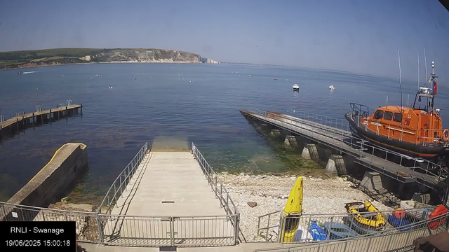 A calm seaside scene with a pier extending into the water, adjacent to a small dock with an orange rescue boat moored on the right. Several small boats are anchored further out in the water. There's a yellow kayak and some small play boats on the pebbled beach area below the dock, along with a surfboard. In the background, a cliffside landscape stretches across the horizon under a clear blue sky. The timestamp indicates the date and time as June 19, 2025, at 3:00 PM.