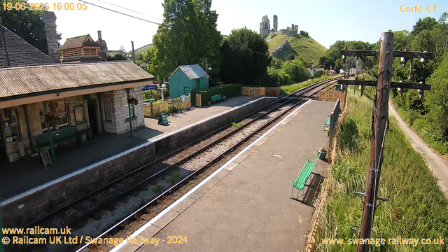 A small train station platform with a building on the left, featuring stone walls and a corrugated roof. There are green benches along the platform and a small green shed behind. The railway tracks run through the center, curving toward a hilltop with ruins of an ancient castle in the background. On the right side, there are wooden utility poles with electrical wires, a narrow footpath, and lush green trees and grass surrounding the station area. The sky is clear and blue.