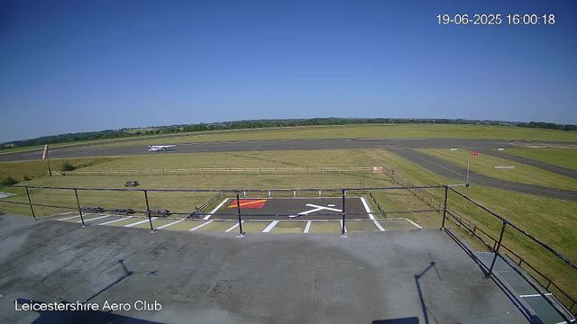 A view from a webcam at Leicesterhire Aero Club shows a clear day with a bright blue sky. In the foreground, there is a small helipad marked with a white helicopter symbol and a red square, surrounded by a black fence. Below the helipad, the words "Leicestershire Aero Club" are visible. Beyond the fence, there is a grassy area, a picnic table, and a runway in the distance. A small white airplane is parked on the runway, and the landscape stretches out to green fields with a few trees under the horizon. The timestamp in the upper right corner indicates the date as 19th June 2025 and the time as 16:00:18.