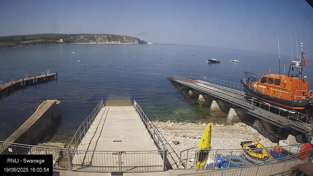 A scenic view of a harbor with calm water and several boats anchored in the distance. In the foreground, there is a concrete dock with metal railings leading into the water. To the right, an orange rescue or emergency boat is moored on a floating platform, and nearby are yellow kayaks, small boats, and some equipment. The background features a landmass with green grass and cliffs under a clear blue sky.