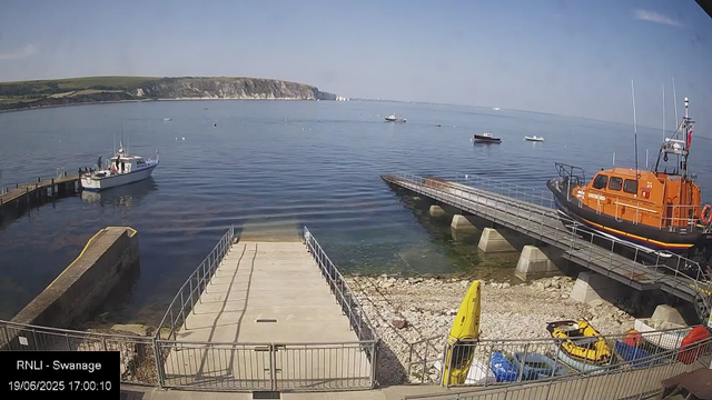 A scenic harbor scene with a few boats floating on calm water, a rocky shoreline in the background, and a clear blue sky. In the foreground, there is a concrete dock with metal railings, and a yellow kayak and some small vehicles parked nearby. An orange rescue boat is docked on the right side, and a few small boats are moored along the shoreline and further out in the water. The overall setting appears peaceful and sunny.