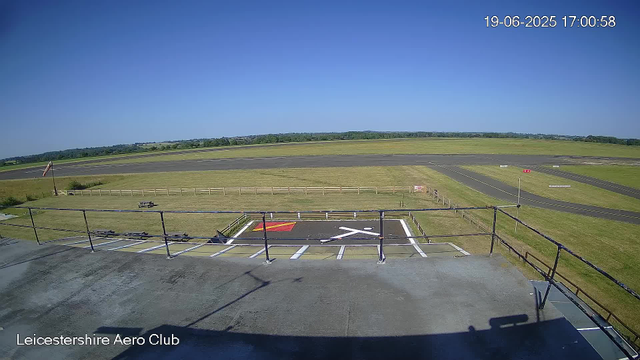 The image shows an empty airport runway and taxiway under a clear blue sky, with a grassy field stretching into the distance. The photo is taken from a camera mounted on a building at Leicestershire Aero Club, as indicated by the label in the bottom left corner. The timestamp in the top right corner reads 19-06-2025 17:00:58. There are some small markings and signs on the ground near the camera, and a fence and picnic table are visible in the foreground. The airport appears quiet with no aircraft present.