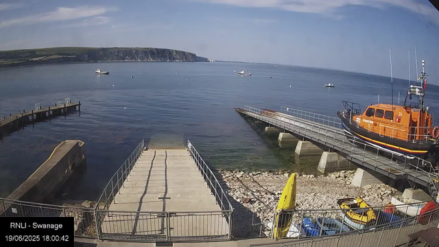 A seaside scene with a calm body of water, several small boats floating, and a rugged cliff in the distance. There are two piers or docks extending into the water, one with a yellow kayak or small boat on the rocky shore. A bright orange lifeboat is docked on the right side, and at the bottom, there are a yellow and a blue kayak along with some parked vehicles. The sky is clear with a few wispy clouds. The timestamp shows the date as June 19, 2025, and the time as 18:00:42.