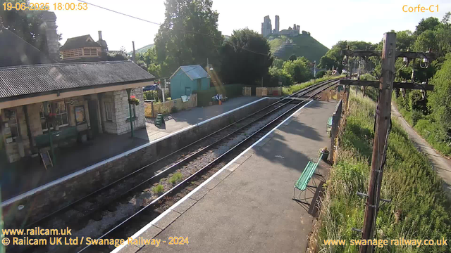 A small railway station platform bathed in late afternoon sunlight. There are two green benches, a wooden utility pole with wires, and a small building with a corrugated metal roof on the left side. In the background, railway tracks extend toward a hill with ruins or an old stone structure atop it. A narrow path runs along the right side of the tracks, bordered by greenery. The timestamp indicates the date and time as June 19, 2025, at 6:00:53 PM.