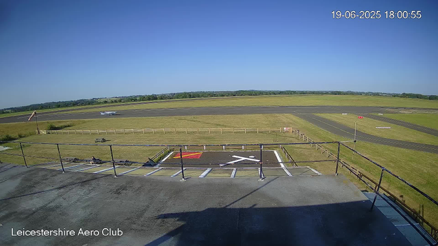 A clear, sunny day at Leicesterhire Aero Club with a blue sky overhead. In the foreground, there is a small designated parking area with white lines, a red marking, and a white arrow painted on the asphalt. A metal railing runs along the edge of the parking area. Beyond the railing, a grassy field surrounds the runway, which stretches horizontally across the image. An aircraft is parked on the runway in the distance, and the landscape extends to a distant tree line on the horizon. The timestamp in the upper right corner reads 19-06-2025 18:00:55.