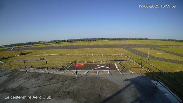 An empty tarmac area at Leicestershire Aero Club with a safety barrier, a small runway section, and a windsock on a pole to the left. The scene is open with grass fields, runway strips, and a clear blue sky in the background. The timestamp reads June 19, 2025, at 6:59 PM.