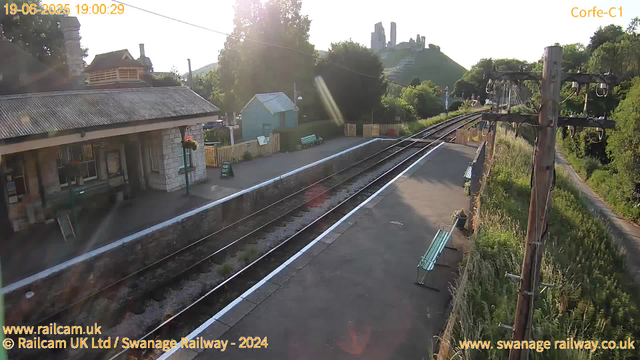 A quiet railway station platform is depicted with a small stone building on the left, featuring bench seating and flower baskets hanging on a lamp post. The train tracks run through the center, curving slightly to the right and disappearing into the distance. There are a few benches along the platform, and a sign indicating "WAY OUT" near the building. On the right side, there are tall wooden railway poles with wires, and greenery and trees along the edge of the platform. In the background, a hill with ruins or a castle is visible, and the sun appears to be setting, casting a warm glow over the scene. The date and time are shown as 19-06-2025 at 19:00:29.