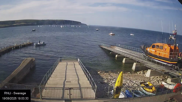 A scenic view of a harbor with several small boats floating on calm water, a large orange rescue boat on a dock, a pedestrian ramp leading into the water, and a rocky shoreline. In the background, there are green cliffs under a partly cloudy sky. The time and date are visible in the bottom left corner.