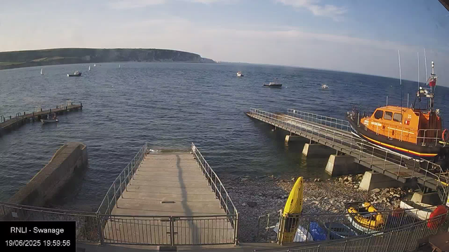 A wide view of a harbor with a dock extending into the water, boats anchored in the sea, and a bright orange rescue or coast guard boat moored to the right. The sky is partly cloudy with a distant landmass or cliffs visible on the horizon. There are some small boats near the dock, and a few boats floating further in the water. The scene appears to be during late afternoon or early evening.