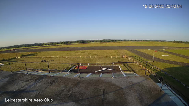 An aerial view of Leicesterhire Aero Club showing a clear sky, the runway in the distance, a small helipad in the foreground with a white letter 'X', a flagpole with a Union Jack flag, a fence surrounding the area, and some grassy fields surrounding the club.