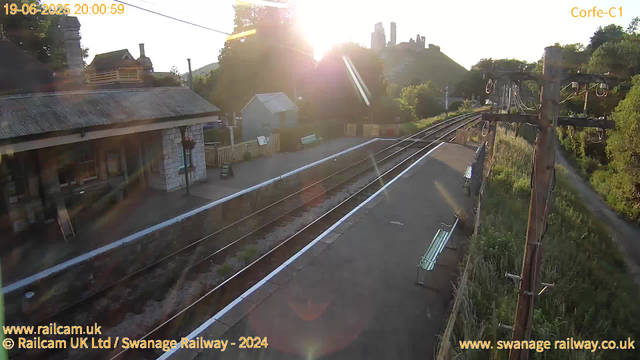 A railway station platform is shown at sunset, with the sun low in the sky creating a bright glare. The platform has benches and a small building with a corrugated metal roof. Overhead electric lines run along the right side, supported by tall wooden poles. A narrow walking path runs adjacent to the station, bordered by trees and greenery. In the background, there is a house on a small hill and a castle ruin visible against the sky. The scene appears calm, with no trains or people present.