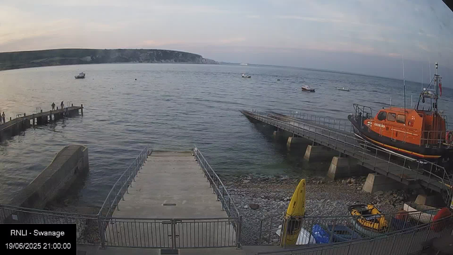 A view of the harbor at sunset, with boats floating on calm water, a wooden pier extending into the sea on the left, and a metal pier with a rescue boat on the right. There are a few people standing on the pier on the left. A large orange rescue boat is docked on the right side of the metal pier. The sky is partly cloudy with soft hues of pink and blue, and a distant shoreline with cliffs can be seen in the background.