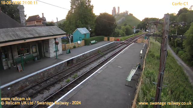 A small railway station platform during daytime with a stone building featuring a sloped roof on the left. There are green benches, potted plants, and a "WAY OUT" sign on the platform. Adjacent to the platform are multiple railway tracks with some grass growing between them. To the right, there are wooden utility poles with electrical wires and a pathway runs parallel to the railway line, bordered by tall grass and bushes. In the background, a hill with ruins or castle-like structures on top is visible, along with trees and greenery. The date and time are displayed at the top left corner as "19-06-2025 21:00:11".