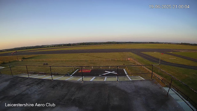 An empty parking area with a helipad marked with a helicopter symbol in the foreground. Beyond the parking area, there is a fenced grassy field and a paved track, all set against a wide open landscape under a clear sky at dusk. The timestamp in the upper right corner shows June 19, 2025, at 9:00 PM. The lower left corner has the text "Leicestershire Aero Club."