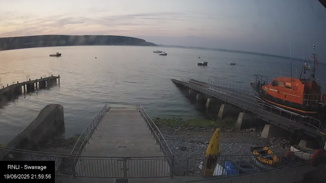 A view of a calm harbor at sunset with several boats floating on the water. There is a small dock extending into the water on the left, and a larger orange rescue boat on a ramp on the right. In the lower foreground, there are some parked vehicles and a yellow kayak. The sky is partly cloudy with soft pastel colors, and a distant shoreline with cliffs is visible across the water.