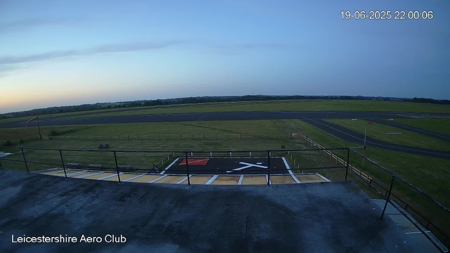 A view from a webcam at Leicestershire Aero Club showing an empty helicopter landing pad with a black and yellow surface, surrounded by green grass and a runway in the distance. The sky is clear with a slight gradient from light blue to darker blue, and the date and time are displayed in the top right corner as "19-06-2025 22:00:06."