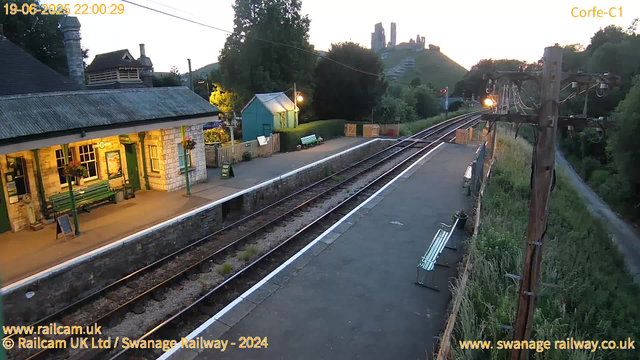 A small train station platform is shown during dusk or early evening, with a building on the left that has hanging flower baskets and outdoor benches. There are two empty benches along the platform. Overhead, part of an electrical pole and wires can be seen. The railway tracks run through the center of the image, with a scenic hill and castle ruins visible in the background on the right. The scene is illuminated by streetlights and the natural fading light of sunset. The image also contains website credits at the bottom.