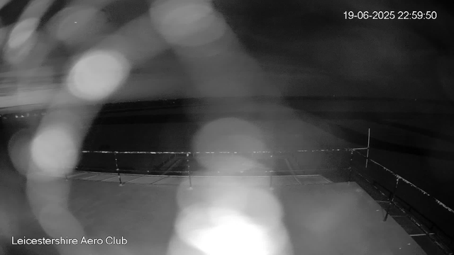 A black and white image taken at night shows a view from a webcam at Leicesterhire Aero Club. The scene appears to be an open outdoor space with a flat surface, possibly a runway or tarmac, enclosed by a railing or fence. There are several out-of-focus bright spots (lens flares or reflections) scattered across the image. The timestamp in the top right corner reads "19-06-2025 22:59:50," and the bottom left corner contains the text "Leicestershire Aero Club." The overall scene is dark with limited visibility of distant objects.