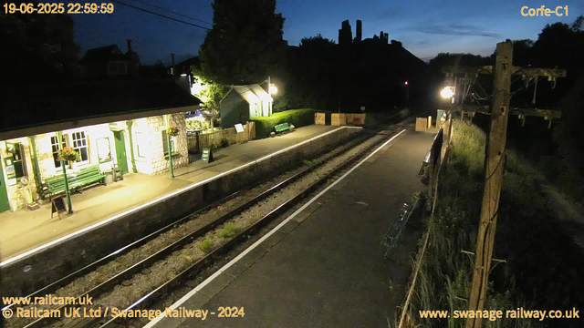 A nighttime scene at a small railway station shows a platform illuminated by yellowish lights. The station building, on the left side, has green doors and window frames, with flower baskets hanging outside. There are benches, a small signboard, and potted plants near the building. The railway tracks run through the center of the image, extending from the foreground into the distance. On the right side, there is a pathway separated from the tracks by a fence, with a few benches and a wooden fence in the background. The sky is dark with a hint of blue, indicating it's shortly after sunset or pre-dawn. There are power poles and lines along the right side. The timestamp in the top left corner reads 19-06-2025 22:59:59.