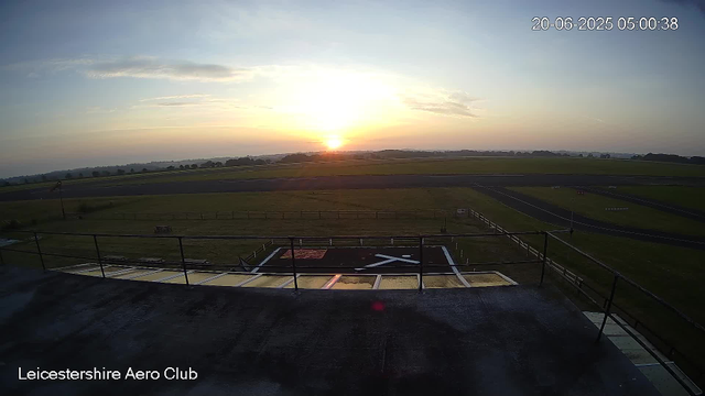 A view from a webcam at Leicesterhire Aero Club showing a runway and open fields under a sunrise or sunset sky with scattered clouds. The timestamp indicates the date and time as June 20, 2025, 5:00 AM. The foreground includes a helipad with a white helicopter symbol, and the bottom left corner displays the text "Leicestershire Aero Club."