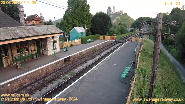 A small railway station platform with a building on the left, featuring a green bench and hanging flower baskets. There are a few benches along the platform, a "Way Out" sign, and a green shed further back. A track runs through the center, with lush green trees and a hill with ruins at the top in the background. Power lines and poles run along the platform's right side, with overgrown grass and a narrow path beyond. The sky is clear, indicating early morning or late evening.