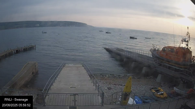 A wide view of a harbor at sunrise showing a concrete pier and a boat ramp in the foreground, a rescue boat on the right side, and a small yellow boat along with some blue containers at the bottom. Several small boats are floating on the calm water, and a large landmass with cliffs is visible in the background under a cloudy sky.