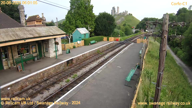 A small train station platform with a stone building featuring a tiled roof, green benches, and flower baskets hanging from the building. There is a green "Way Out" sign on the platform, a small green shed, and a yellow fence near the edge of the platform. Train tracks run alongside the platform, with overhead electrical wires supported by wooden poles. In the background, a lush green hill with a castle ruin on top and a steep staircase leading up the hill. The scene is calm and appears to be early morning, as indicated by the timestamp.