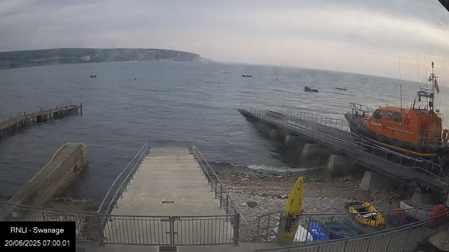 A view of a harbor with a concrete pier extending into the water, on which an orange rescue boat is moored. Several small boats are floating on the water in the distance. The sky is overcast, and there are cliffs visible on the horizon across the bay. Near the bottom of the image, there are some yellow and black safety signs and equipment. The timestamp indicates the date and time as 20/06/2025 at 07:00:01.