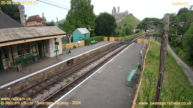 A small train station platform with a stone building on the left, featuring hanging flower baskets and green benches. There are yellow fences, potted plants, and signs near the platform. Multiple railway tracks run through the scene, and overhead electrical lines are supported by wooden poles on the right side. In the background, a grassy hill with ruins or an old castle is visible, and trees surround the area. The scene is captured in clear daylight.