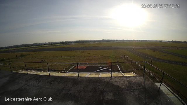A view from a webcam at Leicestershire Aero Club shows a sunny sky with the sun low on the horizon. The scene includes a grassy airfield with taxiways and runways in the distance. The foreground features a platform with a railing, and there are some markings on the ground below. The timestamp in the top right corner indicates the date and time as June 20, 2025, at 7:00:31 AM.