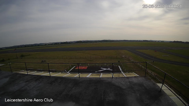 A view from a webcam overlooking the Leicesterhire Aero Club's runway and surrounding grassy landscape. The sky is mostly cloudy with some sunlight breaking through, and the timestamp indicates it is morning on June 20, 2025. The foreground shows a platform with safety barriers and markings, while the background features open fields and the runway extending into the distance.