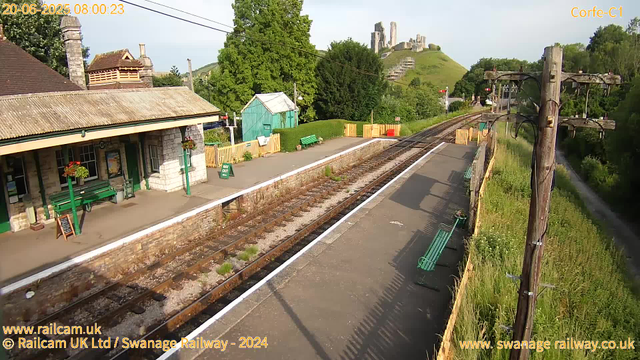 A small rural railway station is shown under bright daylight. There is a single platform with green benches, a small green building, and a train station building made of stone with a tiled roof and flower baskets hanging outside. Railway tracks run through the scene, bordered by a low stone wall. Tall development poles with electrical wires extend along the right side of the platform. In the background, there are lush green trees, a small hill with what appears to be ruins or old structures on top. The scene is peaceful, with shadows indicating the sun is shining. The image includes some branding and website information at the bottom.