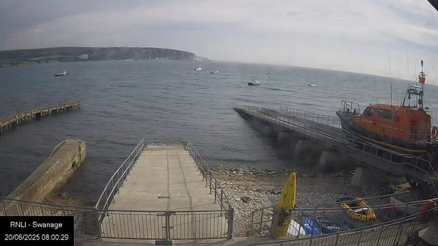 A view of a small harbor with a concrete pier extending into the water. Several boats are floating on the water, and a larger orange rescue boat is docked on the right side on a slipway. In the foreground, there are a few parked vehicles and a yellow caution sign on the rocky shore. The sky is partly cloudy, and distant cliffs rise along the horizon. The timestamp indicates the photo was taken on June 20, 2025, at 8:00 AM.