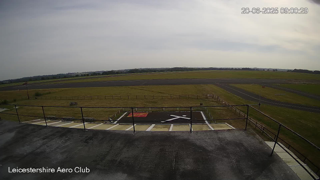 An outdoor view from a webcam at Leicesterhire Aero Club shows a distant runway and grassy fields under a cloudy sky. In the foreground, there is a helipad with a white H marked on a black surface, surrounded by a railing. The timestamp in the upper right corner indicates the date and time as June 20, 2025, at 09:00:29.
