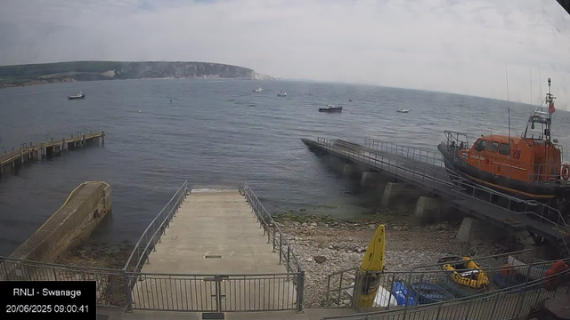 A view of a harbor with several boats on the water, a dock extending into the sea, and a bright orange rescue or patrol boat on a slipway to the right. There is a rocky shoreline with a yellow caution sign and some small vehicles near the bottom. In the background, there are green rolling hills and cliffs under a cloudy sky. The timestamp reads "20/06/2025 09:00:41."