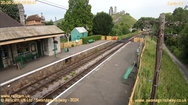 A small train station platform with a building on the left side, featuring stone walls and a brown tiled roof. There are green benches, a green "Way Out" sign, and potted plants on the platform. Two railway tracks run through the scene, with a set of tracks curving towards a castle situated on a hill in the background. The platform is enclosed by a yellow wooden fence, and there are green benches along the right side, with some plants in pots. To the right, there are wooden utility poles with wires stretching across the scene. Behind the station, there is a lush green landscape with trees and a path running parallel to the railway. At the top, the date and time are displayed as "20-06-2025 09:00:47," and the castle on the hill suggests a historic or scenic location.
