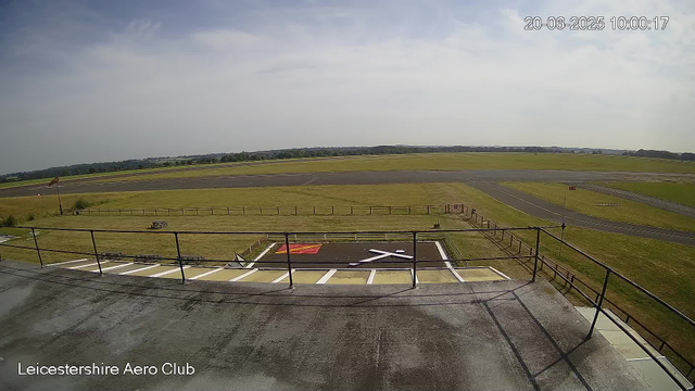 A view from a webcam at Leicesterhire Aero Club showing an airport runway and grassy fields under a partly cloudy sky. The timestamp in the top right corner reads "20-06-2025 10:00:17." In the foreground, there is a helipad marked with a large white "H" inside a black square, and a fencing area surrounds the helipad. The bottom left corner displays the text "Leicestershire Aero Club." The scene appears calm and empty, with no aircraft or people visible.