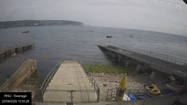 A view of a harbor with a small pier extending into the water, which is calm with slight ripples. Several boats are anchored in the distance, and a cliffside is visible on the horizon under a cloudy sky. In the foreground, there is a concrete area with a railing, a small yellow boat, yellow safety equipment, and some containers. A yellow caution sign is positioned on the ground near the railing.