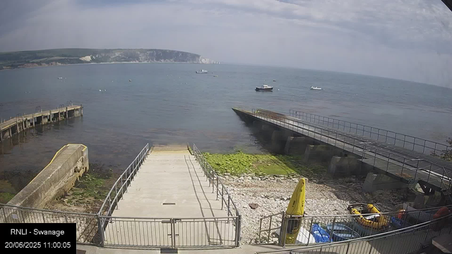 A view of a seaside area with a small pier leading into the water. Several boats are anchored in the calm sea, and there is a distant coastline with cliffs and land. The sky is partly cloudy, and the time and date are displayed in the bottom left corner as "20/06/2025 11:00:05".