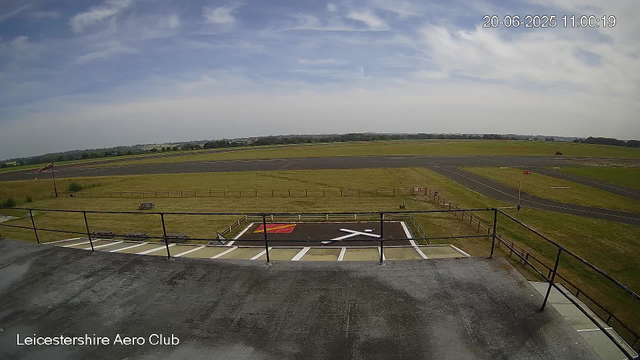 An outdoor view from a webcam at Leicestershire Aero Club showing a runway and grassy areas under a partly cloudy sky. The image includes a parked helicopter landing pad in the foreground, with markings and a railing, and a windsock on a pole to the left. In the distance, there are fields, trees, and a small vehicle on the runway. The timestamp in the top right corner reads "20-06-2025 11:00:19."
