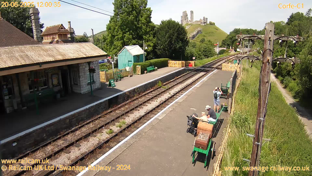 A small train station platform with a stone building on the left, green benches, and a few people sitting or standing. There are railway tracks running parallel along the platform. In the background, a hill with a castle ruin on top, surrounded by lush green trees. An old wooden lamppost with multiple lamps stands on the right side of the platform. The sky is clear, and the scene appears to be sunny. A date and time stamp indicating "20-06-2025 12:00:35" is visible in the upper left corner.
